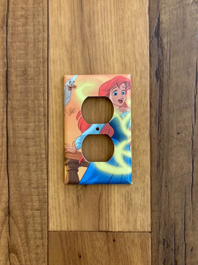 May include: A light switch cover featuring a colorful illustration of Ariel from The Little Mermaid. She is wearing a blue dress and has red hair. A seagull is flying behind her.