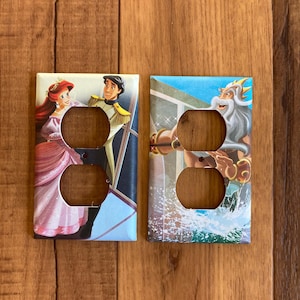 May include: Two decorative light switch covers featuring Disney characters. One shows Ariel and Prince Eric, the other shows King Triton. The covers are rectangular with two outlet holes and are set against a wooden background.