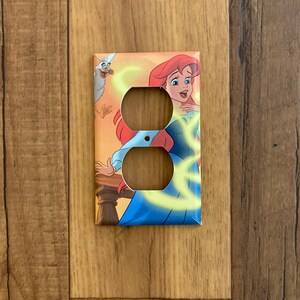 May include: A light brown double outlet cover with a colorful image of Ariel from The Little Mermaid. Ariel is wearing a blue dress and has red hair. She is surrounded by yellow swirls and a seagull.