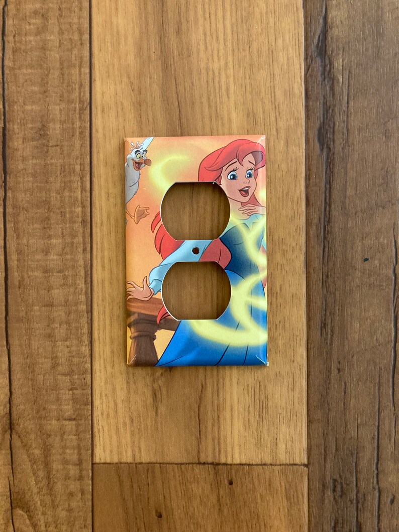 May include: A light brown double outlet cover with a colorful image of Ariel from The Little Mermaid. Ariel is wearing a blue dress and has red hair. She is surrounded by yellow swirls and a seagull.