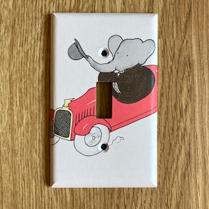 May include: A white light switch cover featuring a cartoon illustration of an elephant in a suit driving a red car. The elephant is wearing a hat and waving. The switch opening is a dark brown rectangle.