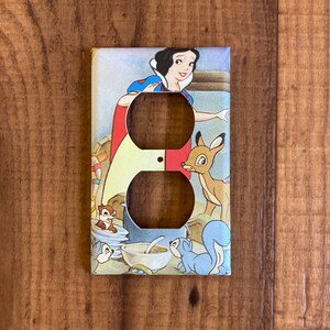 May include: A white light switch cover with a vintage illustration of Snow White and the Seven Dwarfs. Snow White is wearing a blue and red dress and a yellow crown. She is surrounded by woodland animals, including a deer, a squirrel, and a chipmunk.