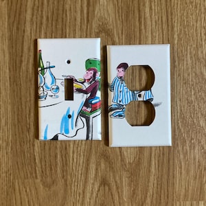 May include: Two white electrical outlet covers with illustrations. One features a monkey at a table with bottles and glasses. The other shows a person in blue and white striped pajamas. The covers are rectangular with screw holes and outlet cutouts.