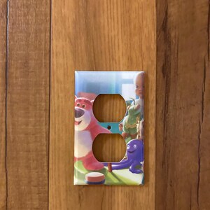 May include: A light blue and white outlet cover with a colorful image of characters from the Toy Story movie franchise. The characters include Woody, Buzz Lightyear, and a purple octopus.