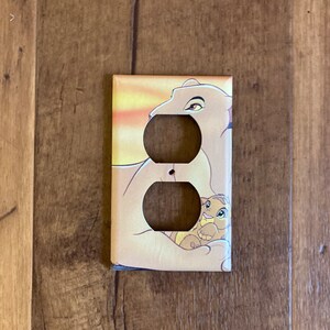 May include: A light brown and yellow light switch cover with a cartoon image of a lioness and a lion cub. The lioness is lying down with her paw over the cub. The background is a sunset.