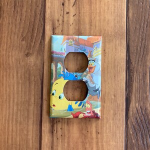 Puede incluir: Una cubierta decorativa para interruptores de luz con personajes de la pel&iacute;cula animada "La Sirenita". La cubierta tiene un dise&ntilde;o colorido con im&aacute;genes de Flounder, Sebasti&aacute;n y otros personajes sobre un fondo de castillo y oc&eacute;ano.