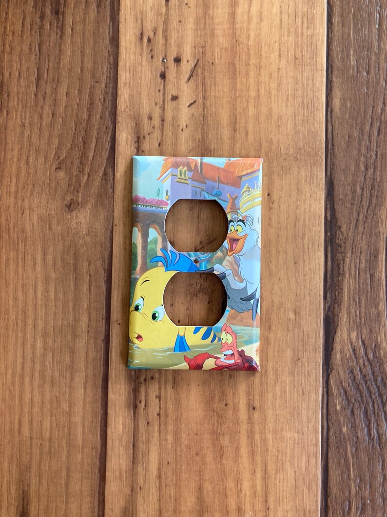 Puede incluir: Una cubierta decorativa para interruptores de luz con personajes de la pel&iacute;cula animada "La Sirenita". La cubierta tiene un dise&ntilde;o colorido con im&aacute;genes de Flounder, Sebasti&aacute;n y otros personajes sobre un fondo de castillo y oc&eacute;ano.