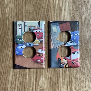 May include: Two rectangular light switch plates featuring colorful graphics from the movie Cars. The left plate displays multiple animated car characters, while the right plate shows a red race car with the number 95 and a blue car in a garage.
