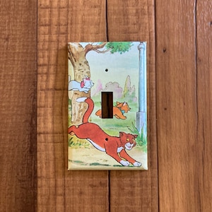 May include: A light switch cover featuring a cartoon illustration of three cats from the Disney movie The Aristocats. The cats are running through a green grassy area with a tree and a fence in the background. The switch cover is white with a light brown border.