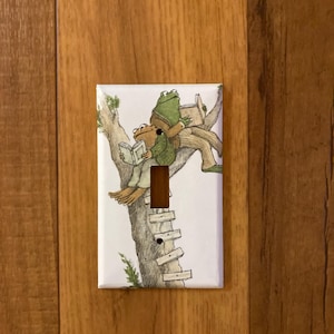 May include: White light switch cover featuring a whimsical illustration of two frogs reading books in a tree. The frogs are wearing jackets and sitting on a tree branch. The switch plate has a brown toggle switch.