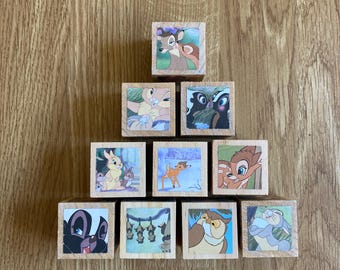 May include: Nine wooden blocks with different Disney characters from Bambi, including Bambi, Thumper, Flower, and the skunk. The blocks are arranged in a pyramid shape.