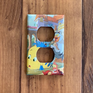 Puede incluir: Cubierta decorativa para interruptor de luz con una colorida ilustraci&oacute;n de personajes de La Sirenita. La cubierta presenta a Flounder, Sebasti&aacute;n y Scuttle con un castillo de fondo. Tiene dos agujeros para los enchufes.