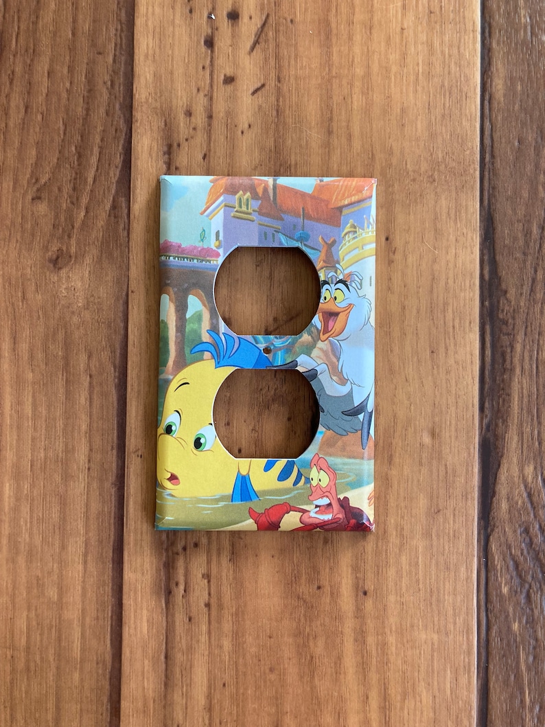 Puede incluir: Cubierta decorativa para interruptor de luz con una colorida ilustraci&oacute;n de personajes de La Sirenita. La cubierta presenta a Flounder, Sebasti&aacute;n y Scuttle con un castillo de fondo. Tiene dos agujeros para los enchufes.