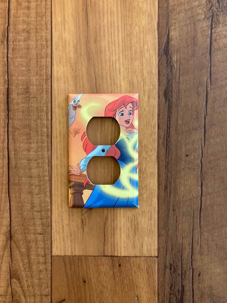 May include: A light switch cover featuring Ariel from Disney's The Little Mermaid. The cover is decorated with a colorful illustration of Ariel, a seagull, and swirling yellow light.