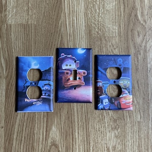 May include: Three rectangular light switch and outlet covers featuring colorful graphics from the movie Cars. The left cover has two circular openings, the center has one toggle switch opening, and the right has two circular openings.