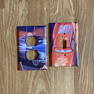 May include: Two decorative light switch covers featuring characters from the animated movie Cars. One cover depicts Sheriff, a blue police car, and the other features Lightning McQueen, a red race car. Both have cutouts for switches.