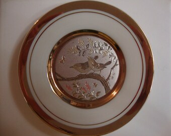 Könnte beinhalten: Ein weißer Porzellanteller mit goldfarbenem Rand und einem kleineren, runden Teller in der Mitte. Der kleinere Teller zeigt ein silberfarbenes Design mit zwei Vögeln, die auf einem Zweig mit Blättern und Blumen sitzen. Das Design ist von einem goldenen Rand umgeben.