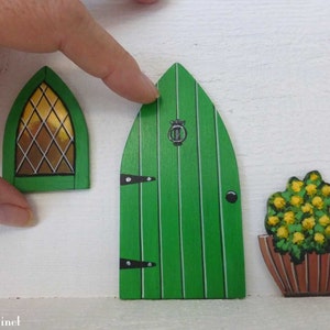 Op de afbeelding: Een set van drie houten decoraties voor een feeënhuis. De set bevat een groene deur met een zwarte deurknop, een groen raam met een gouden glas-in-lood ontwerp en een bruine bloempot met een gele bloem.