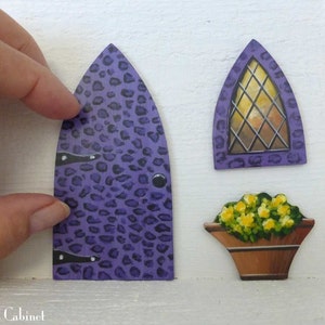 Op de afbeelding: Drie houten uitsnijdingen geschilderd in een fantasierijke stijl. Een paarse deur met een luipaardprint, een paars raam met een ruitvormig glas en een bruine bloempot met gele bloemen.