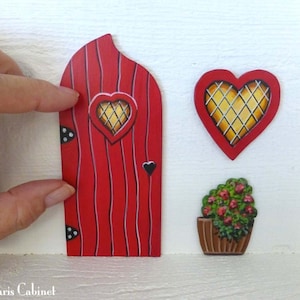 Può includere: Una porta di fata in legno rosso con una finestra a forma di cuore e pois neri. C'è una finestra a forma di cuore con un motivo a griglia dorata e una pianta in vaso con fiori rossi.