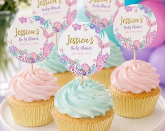 May include: A white cake stand holds cupcakes with pink and blue frosting. Each cupcake has a decorative topper with the text "Jessica's Baby Shower July 15th, 20xx" and a mermaid tail design. The cupcakes are in gold foil liners.