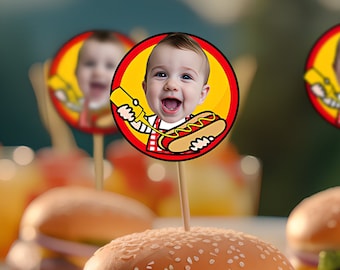 May include: A close-up shot of a cheeseburger on a white plate, with a sesame seed bun, a beef patty, cheese, lettuce, and a pickle. A decorative toothpick with a baby's face holding a hotdog is on top. The background is blurred.
