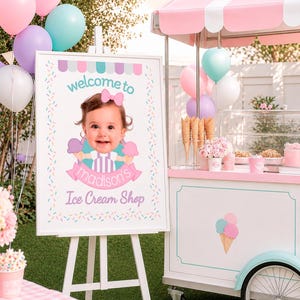 Puede incluir: Un letrero con marco blanco sobre un caballete de madera que muestra la foto de un niño, el texto "welcome to madison's Ice Cream Shop" y un borde de chispas de colores. Al lado hay un carrito de helados blanco y rosa con toldo a rayas.