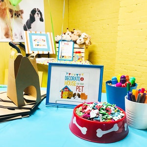 May include: A blue table with a red dog bowl filled with colorful foam shapes. There is a sign that says "Decorate a House for Your Pet" and a cardboard dog house on the table.