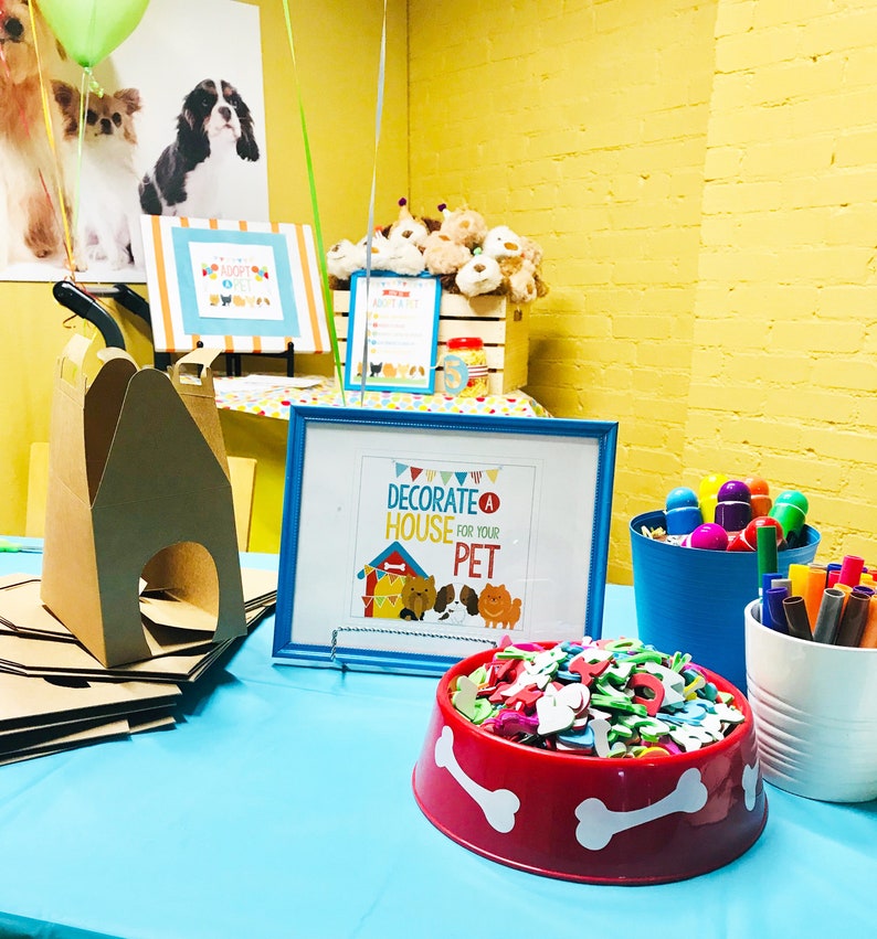 May include: A blue table with a red dog bowl filled with colorful foam shapes. There is a sign that says "Decorate a House for Your Pet" and a cardboard dog house on the table.
