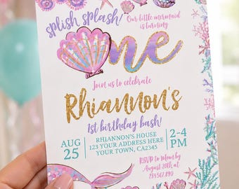May include: A mermaid-themed birthday invitation with the text "splish splash!" and "Our little mermaid is turning ONE." The invitation is decorated with seashells, starfish, and a mermaid tail. The invitation is for Rhiannon's 1st birthday bash on August 25th.