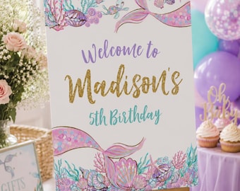 May include: A white sign on a wooden easel with the text "Welcome to Madison's 5th Birthday." The sign is decorated with mermaid tails, seashells, and coral in shades of pink, purple, and teal. Balloons and cupcakes are in the background.