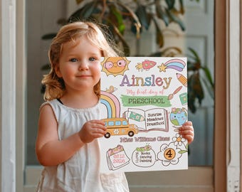 May include: A young child holds a colorful sign with the name "Ainsley" and the text "My first day of Preschool." The sign features illustrations of a school bus, rainbow, pencil, and other school-related graphics. The sign also includes the date 08.12.2026.