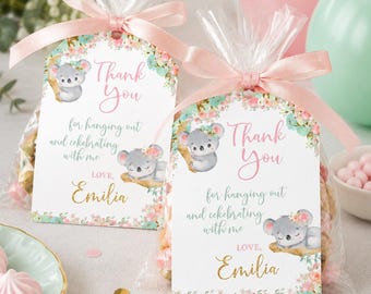 May include: Two clear plastic favor bags filled with treats, each topped with a rectangular tag featuring koala illustrations, floral accents, and the text Thank You for hanging out and celebrating with me LOVE, Emilia, tied with a light pink ribbon.