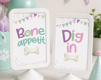May include: Two white rectangular tabletop signs with rounded corners and circular bases. The left sign displays the text "Bone appetit" with a paw print and a bone graphic. The right sign displays the text "Dig in" with a bone graphic.