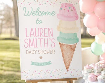 May include: A wooden easel displays a baby shower sign with an ice cream cone illustration. The sign reads "Welcome to Lauren Smith's Baby Shower" in pink and mint green, with the date April 8th, 20XX. Pastel balloons and cupcakes decorate the background.
