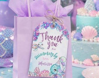 Può includere: Una borsa regalo lilla con un'etichetta che recita "Thank you for swimming by, Love, Marissa". L'etichetta presenta illustrazioni di una tartaruga e una balena. La borsa è riempita di carta velina lilla e ha un manico abbinato.