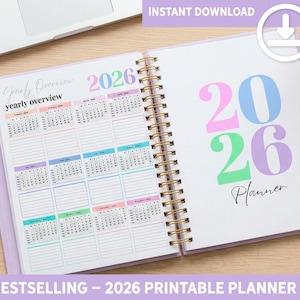 Peut inclure: Un agenda à spirales ouvert, avec une couverture violet clair. La page de gauche affiche un calendrier annuel pour 2026, tandis que la page de droite présente le chiffre "2026" en couleurs pastel. Le texte "Planner" est également visible.