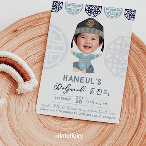 Peut inclure: Une invitation coréenne pour un Doljanchi avec un bébé souriant en tenue traditionnelle. La carte contient du texte en anglais et en coréen, les détails de l'événement et une bordure décorative. Sur une surface en bois.
