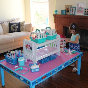 K&ouml;nnte beinhalten: Ein blauer und rosa Spieltisch mit einer rosa Tischdecke und verschiedenen Spielzeug-Medizinutensilien, darunter ein Stethoskop, Bandagen und eine wei&szlig;e Plastik-Tiertransportbox. Ein junges M&auml;dchen steht neben dem Tisch und h&auml;lt eine Spielzeugspritze.
