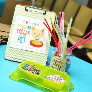 May include: A green plastic dog bowl with two compartments filled with colorful beads. A sign on a clipboard reads "Make a Collar for Your Pet". A jar filled with colorful pipe cleaners sits next to the bowl.