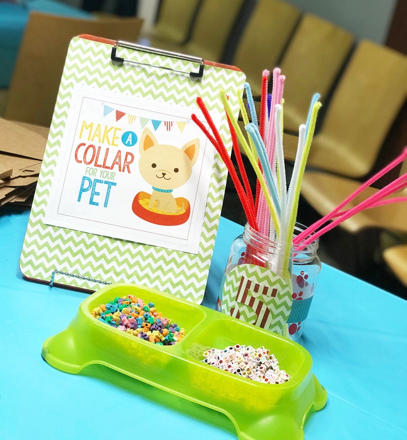 May include: A green plastic dog bowl with two compartments filled with colorful beads. A sign on a clipboard reads "Make a Collar for Your Pet". A jar filled with colorful pipe cleaners sits next to the bowl.
