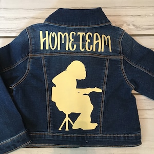 May include: A dark blue denim jacket with the word "HOMETEAM" in a light yellow color across the upper back. A light yellow silhouette of a person playing a guitar is below the text. The jacket has a classic collar and button closures.