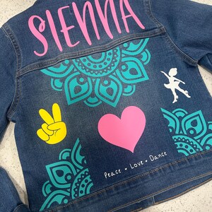 May include: A denim jacket with a teal mandala design, a pink heart, a yellow peace sign, and the name "SIENNA" in pink lettering. The text "Peace Love Dance" is written in white below the heart. A white silhouette of a dancer is on the right side of the jacket.