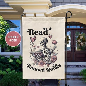 May include: A garden flag with a white background and a skeleton reading a book. The text on the flag reads "Read Banned Books".