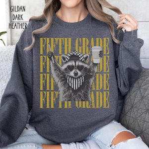 May include: A dark heather gray Gildan sweatshirt featuring a graphic of a raccoon wearing a checkered cap, heart-shaped sunglasses, and a striped bandana. The raccoon holds a pencil and a coffee cup. The text FIFTH GRADE is repeated in gold.