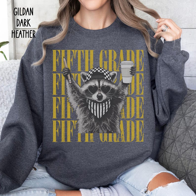 May include: A dark heather gray Gildan sweatshirt featuring a graphic of a raccoon wearing a checkered cap, heart-shaped sunglasses, and a striped bandana. The raccoon holds a pencil and a coffee cup. The text FIFTH GRADE is repeated in gold.