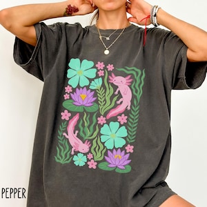 May include: Dark grey t-shirt featuring a colorful graphic of two pink axolotls, surrounded by turquoise flowers, purple water lilies, and green foliage. The word "PEPPER" is printed in white at the bottom left.