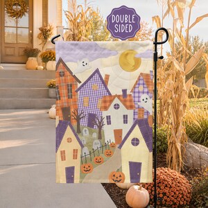 May include: A rectangular garden flag featuring a quilted-style illustration of a neighborhood with houses in orange plaid, purple gingham, and cream. The scene includes a yellow moon, two white ghosts, tombstones, and orange pumpkins. Text reads DOUBLE SIDED.