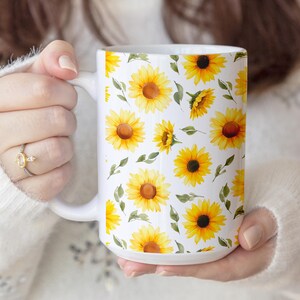 May include: White ceramic mug featuring a repeating pattern of yellow sunflowers with brown centers and green leaves. The mug has a large handle and is held by a person. The floral design covers the entire surface of the mug.