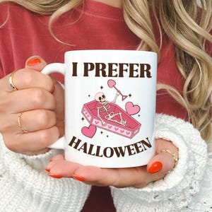 May include: White ceramic coffee mug with a pink and brown illustration of a skeleton in a coffin holding a shovel. The text "I prefer Halloween" is printed on the mug.